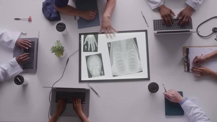 An overhead shot captures six individuals some in lab coats sitting around a white table and actively reviewing several xray films placed on an illuminated screen while working on laptops and taking notes