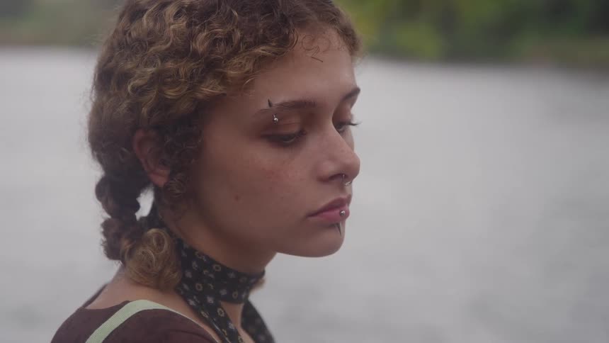 A pensive young woman with facial piercings lowers her gaze standing next to a river