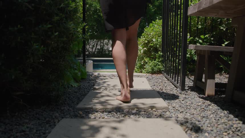 Female legs crossing a walkway and going through a gate on a lawn towards a swimming pool outdoors during day time