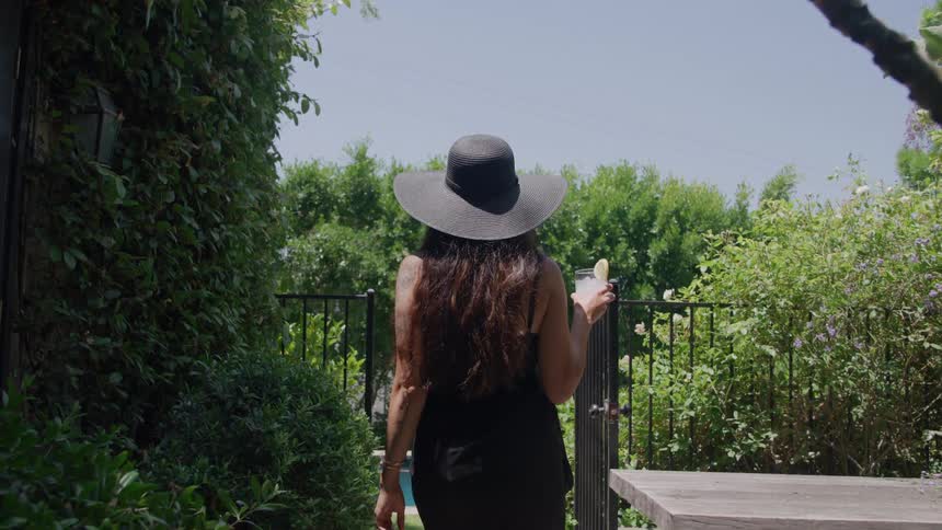 A shot from behind captures a woman in a large black sunhat and dark attire walking away from a table through a lush garden holding a cocktail glass as she approaches a black metal gate