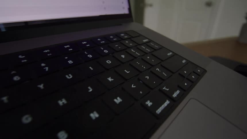 This is an extreme closeup video focusing on the keyboard and trackpad area of a laptop likely a macbook pro due to the key arrangementthe camera pans diagonally across the keyboard revealing the backlit keys in a dimly lit indoor environment