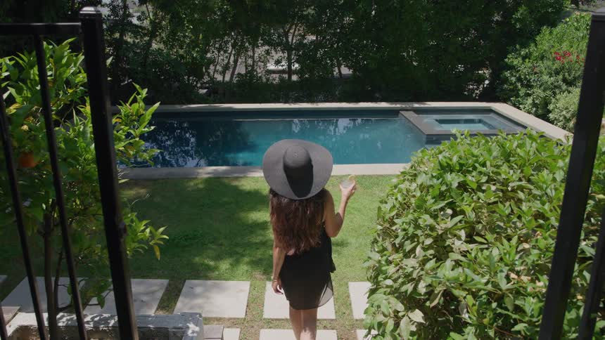 Woman wearing a hat walks through a garden toward a pool