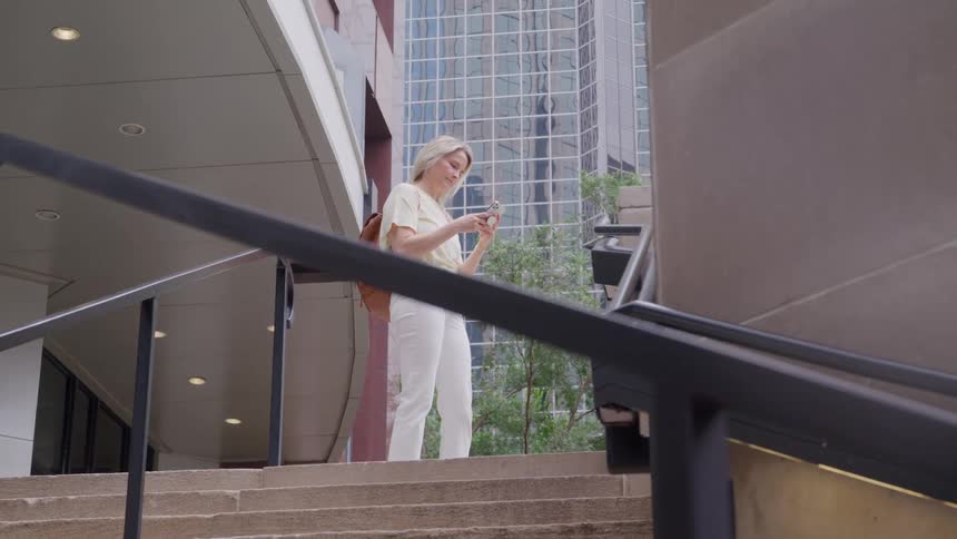 This video shows a young woman standing on a set of outdoor concrete stairs looking at and interacting with her mobile phoneshe is wearing a light yellow tshirt white pants and a light brown backpack with a modern glassfaced skyscraper visible i