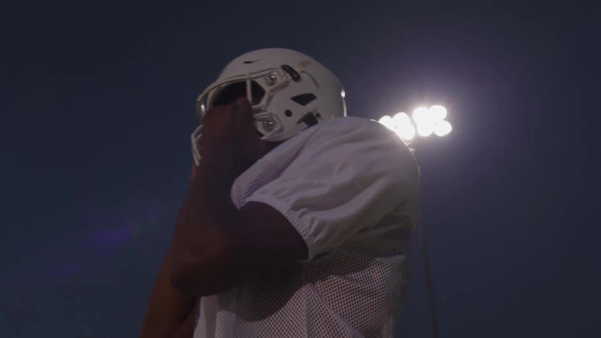 Football player stands on under the stadium lights and puts on his helmet