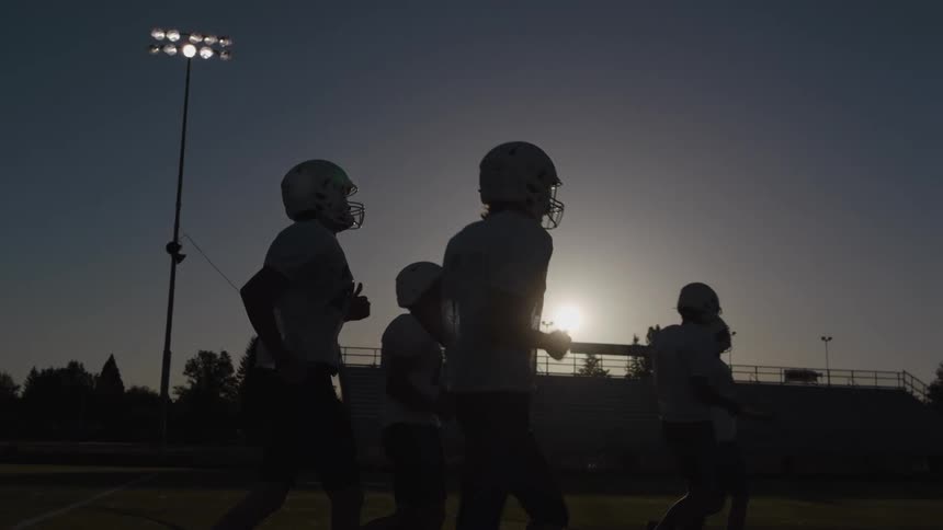 American football players run on the field at dusk with bleachers in the background