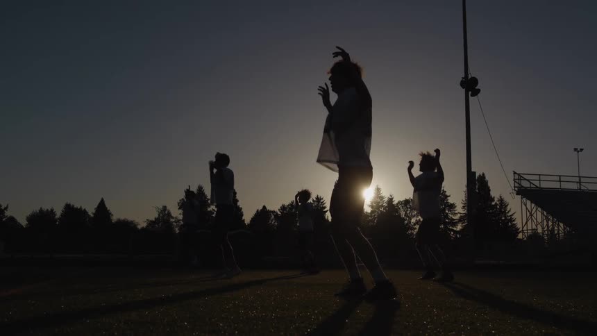 This video is a dramatic lowangle silhouette shot of a group of athletes exercising on a sports field at sunset or sunrisethe athletes likely football players are performing jumping exercises with the deep shadows highlighting their movements agai
