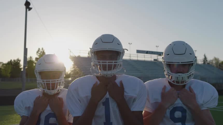 A medium shot captures three football players in white jerseys and helmets standing in a line on a field adjusting their collars as the setting sun shines brightly behind them in the stadium