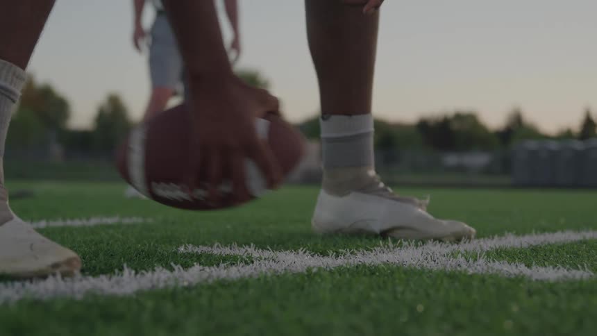 Close up of a black athlete throwing a football in between their legs while playing on an outdoor field at sunset