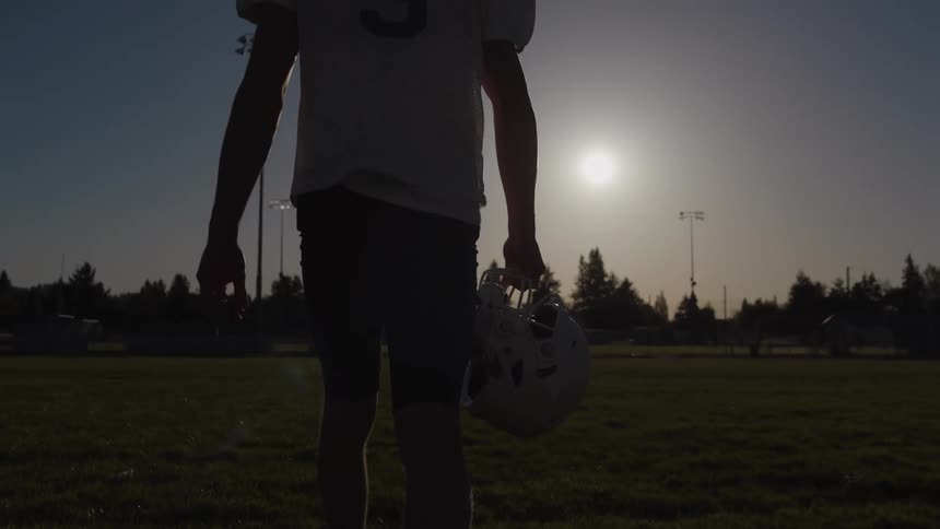 Football player walks across the field carrying his helmet in his hand