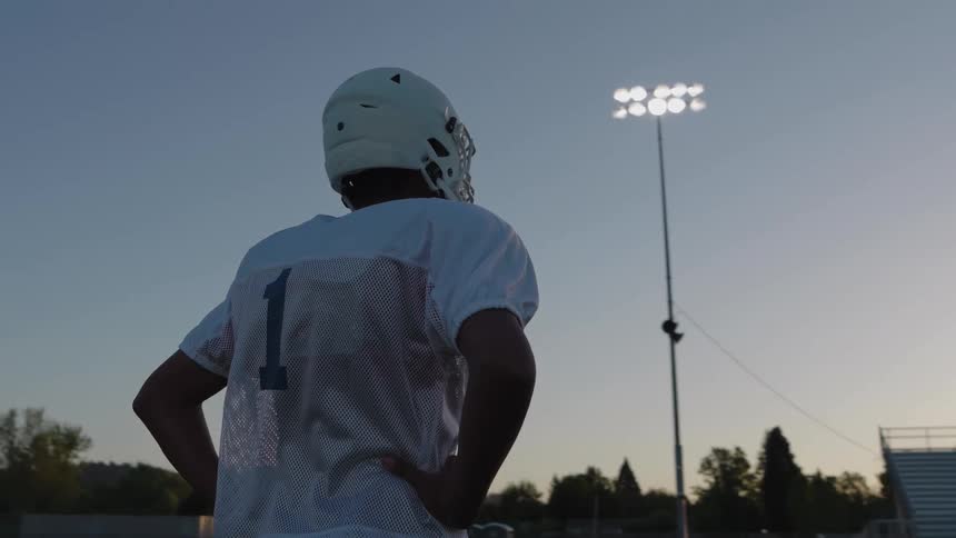 American football player wearing a helmet and number 1 jersey stands on the field in front of the bleachers at dusk