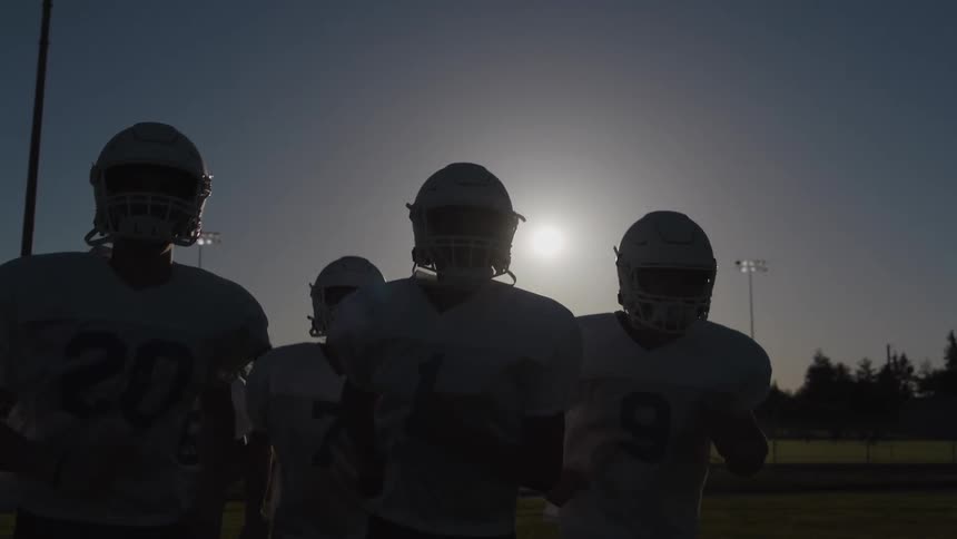 This video features a group of american football players wearing helmets and practice jerseys running slowly toward the camerathe players are heavily backlit by the sun creating a strong silhouette effect that emphasizes their helmets and gear agai