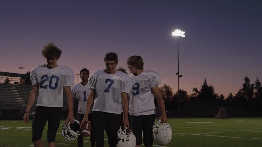 American football players walk across the field at dusk carrying helmets and a ball