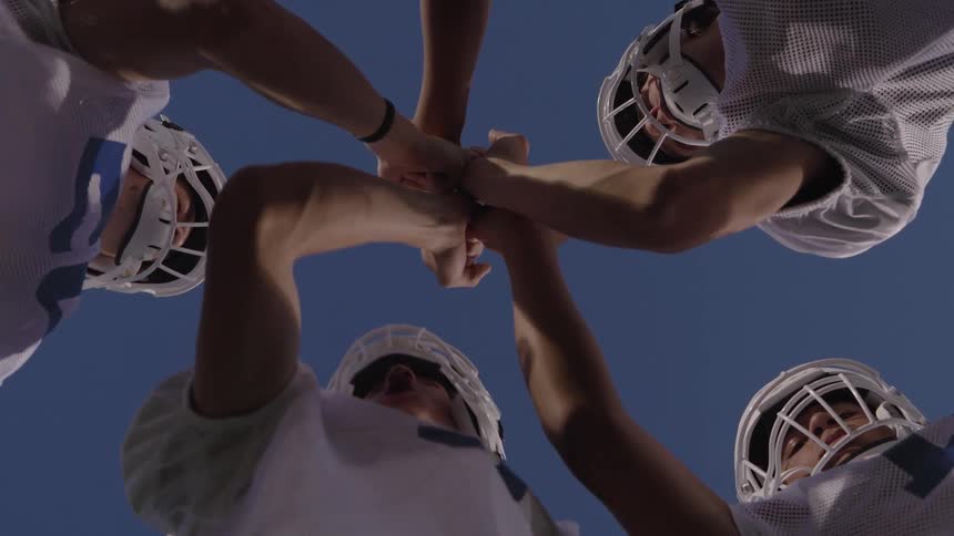 American football players form a circle with their hands joined viewed from below against the sky