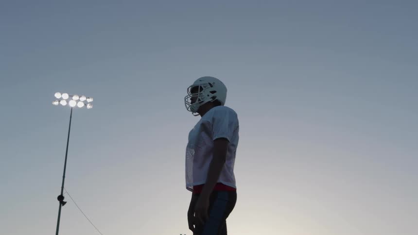 Football player wearing a helmet and practice vest stands on the field at dusk