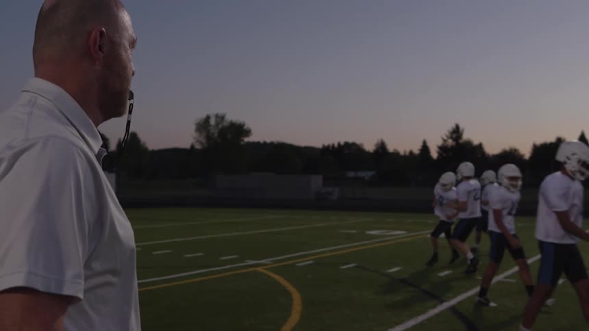 Coach watches american football players walking across the field at dusk