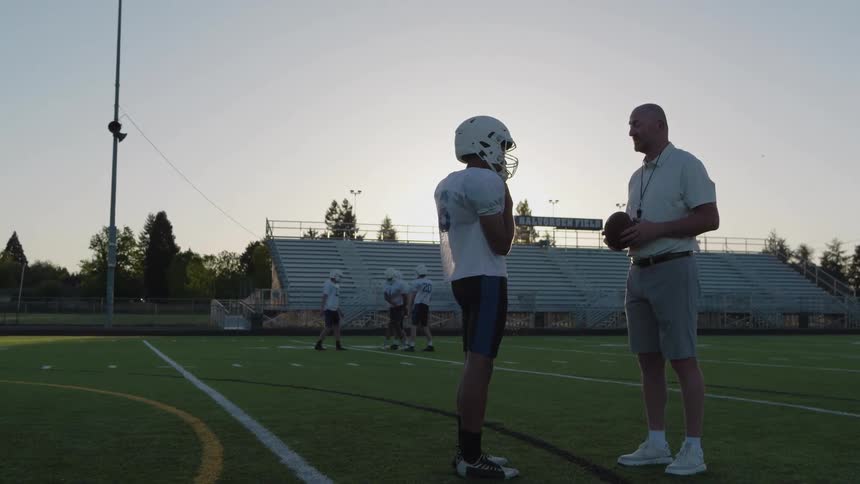 Coach instructs football player during sunset practice on field with bleachers in background