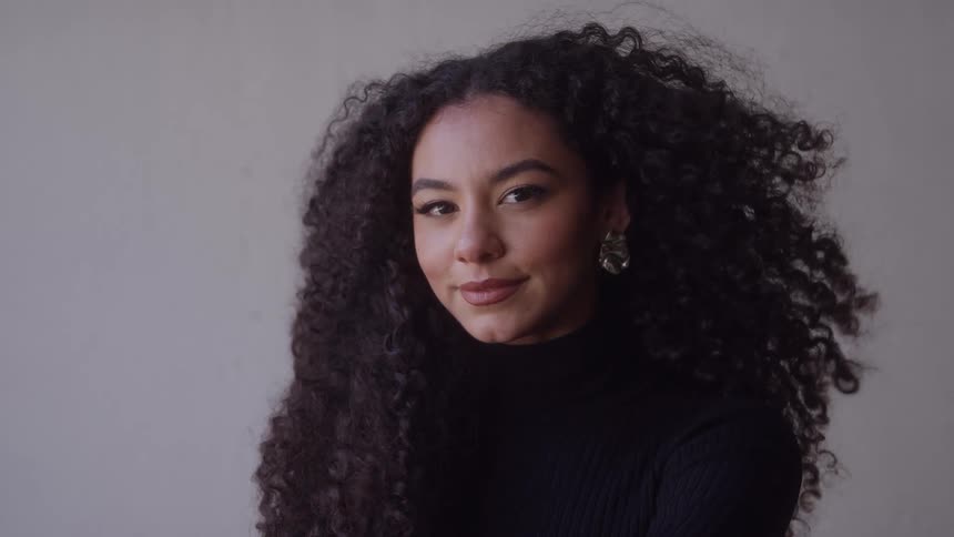 A beautiful woman with voluminous dark curly hair and large earrings stands against a plain background with her eyes closed in a moment of tranquility