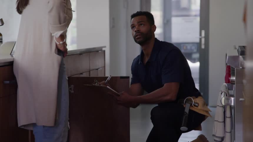 A handyman inspects a kitchen cabinet closely jotting down notes as he listens attentively beside him a woman explains the issue shes been experiencin