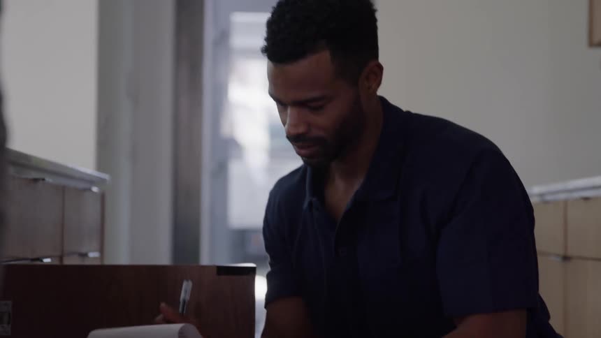 A medium closeup shot focuses on a man in a dark polo shirt appearing to be a delivery person or technician as he handles paperwork and a pen he glances down seemingly reviewing or signing a document related to a nearby piece of furniture or cabinetr