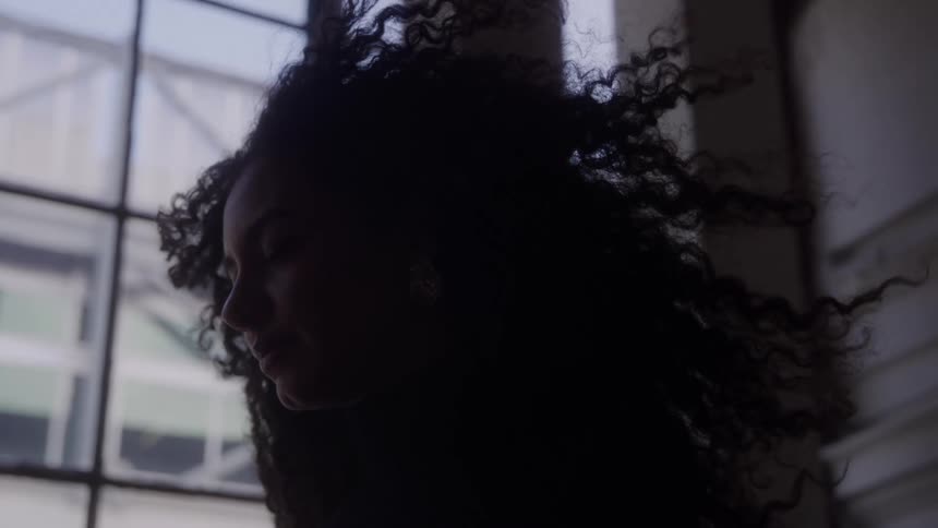 Woman in profile with loose hair stands indoors near a window