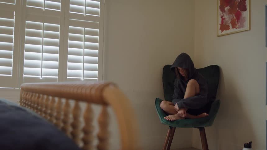 A young person is seen sitting alone curled up in a dark hooded sweatshirt in a teal armchair in the corner of a bedroomthe person is hugging their knees to their chest and appears somber with wooden plantation shutters visible on the window nearby