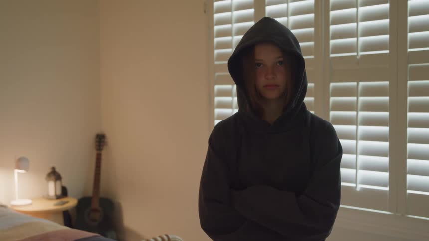 Girl wearing a hooded sweatshirt stands in a bedroom lit by a window with shutters