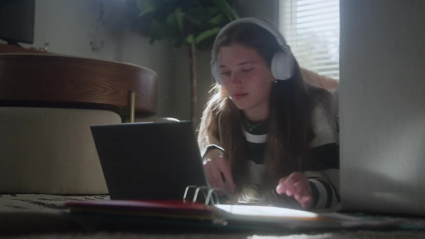 A teenage girl wearing white headphones lies on a cozy carpet concentrating on writing notes with a pencil while referencing information displayed on a small laptop in a softly lit indoor setting