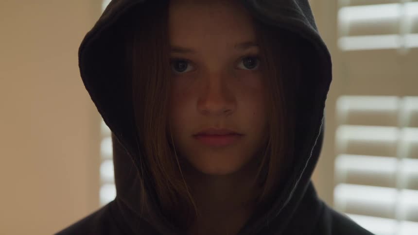 A close view of a young woman wearing a black hoodie with the hood up looking down with her eyes closed against a background of bright window blinds