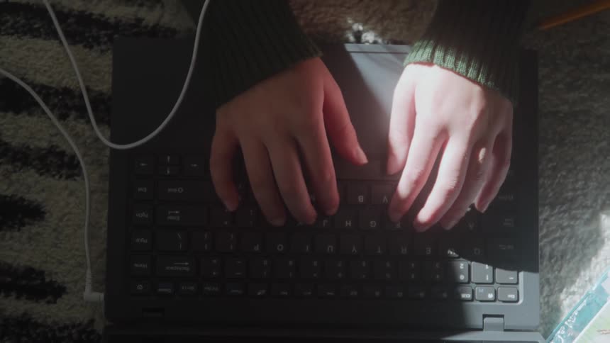 Hands type on a laptop keyboard placed on a rug with a cable connected