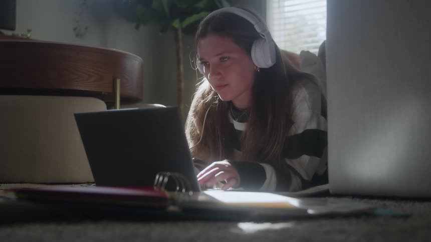 A young woman is lying on the floor wearing headphones and glasses and working intently on a laptop that is propped up in front of hersunlight streams in from a nearby window illuminating the scene and school supplies like a binder and notebooks ar
