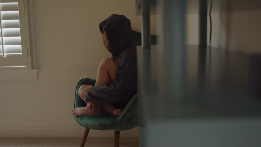 Woman sitting in an armchair wears a hooded sweatshirt and keeps their legs tucked while looking toward the window