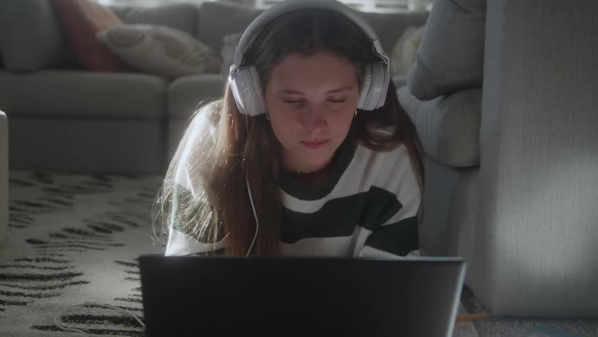 A young woman wearing large white overear headphones and a striped sweater is lying on her stomach on the floor in a living roomshe is focused intently on a laptop screen placed in front of her occasionally looking up with a thoughtful or concentrate