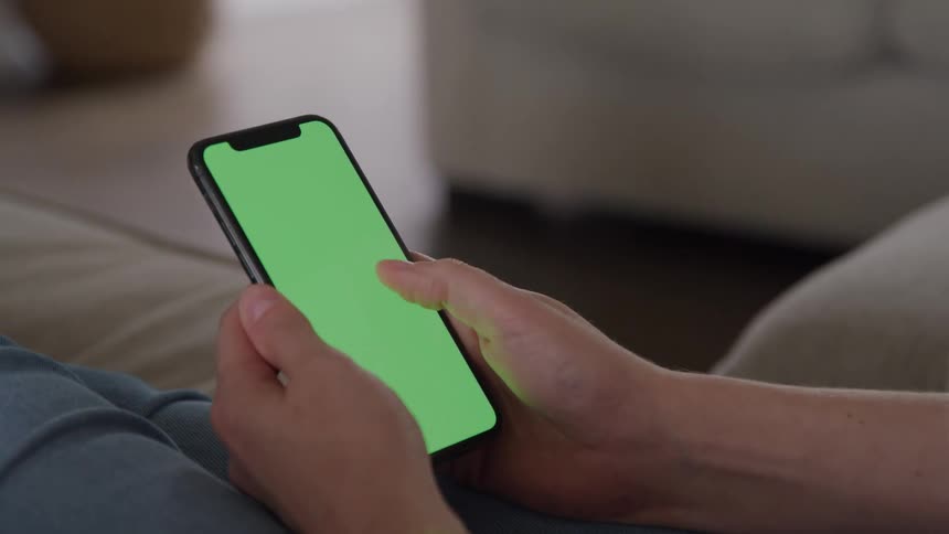 The video shows a closeup of a person sitting down holding a modern smartphone in portrait orientationthe person is using their thumb to scroll or interact with the screen which is replaced with a green screen for digital insertion