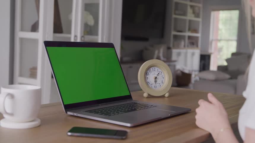 Woman sitting at a table in front of a laptop computer with a green screen as she waves at the computer screen