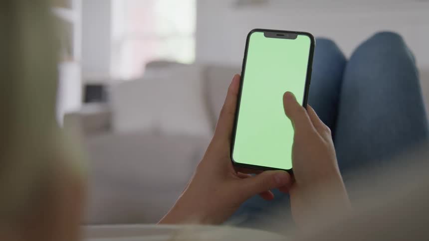 A closeup overtheshoulder shot shows a person reclining on a couch holding and interacting with a smartphone that has a green screen display the person uses their thumb to tap swipe and scroll on the screen several times suggesting they are browsi