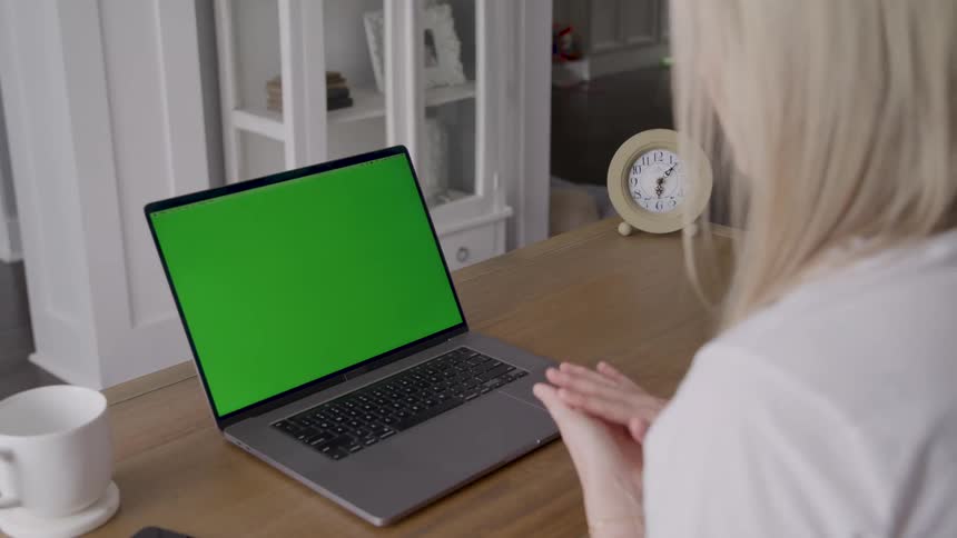 A person with blonde hair is sitting at a wooden desk looking at a laptop with a green screen ready for content replacementthe person is actively engaged using expressive hand gestures as if speaking to someone on a video call or explaining something