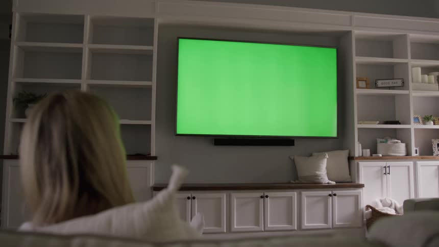 Young adult woman sitting down on a couch pointing a remote control at a television displaying a replaceable screen indoors during day time