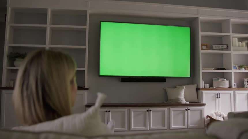A woman watches a television with a green screen mounted on built in white shelving and cabinetry in a contemporary living room