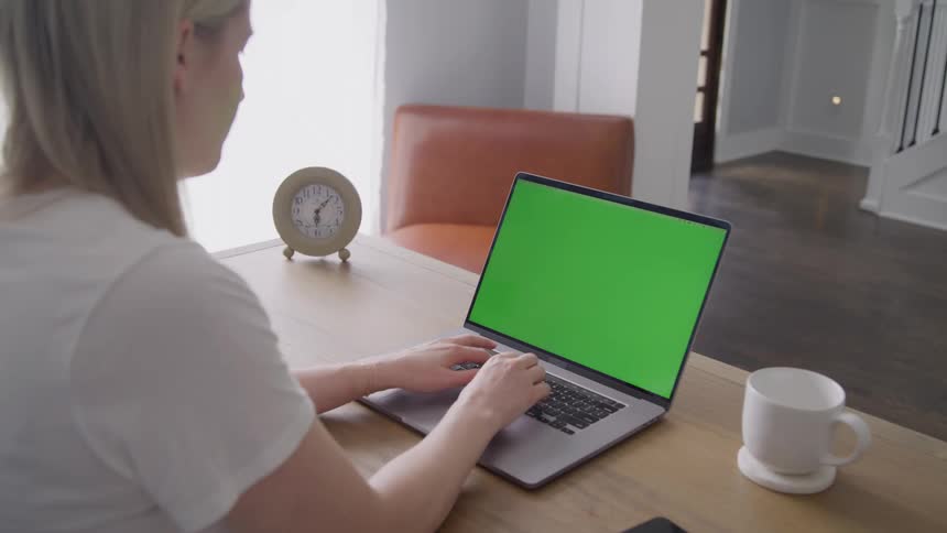 A female sitting at a desk with a clock and a mug is typing on a laptop in a room
