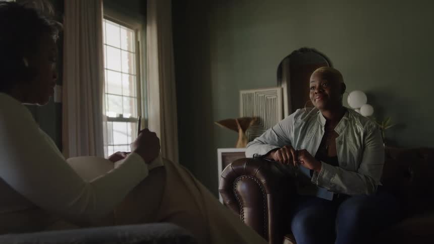 Two black adult females are having a seated conversation in a dimly lit elegantly decorated living room with one smiling and looking forward as another is taking notes