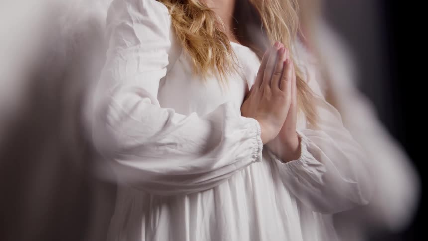 Woman in a white dress holds their hands together in a praying position beside decorative wings