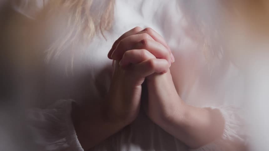 A tightly framed closeup focuses on a persons hands clasped together near their chest in a gesture of prayer or contemplation the scene is softly lit creating a bright slightly blurred halo effect around the persons white clothing and hair