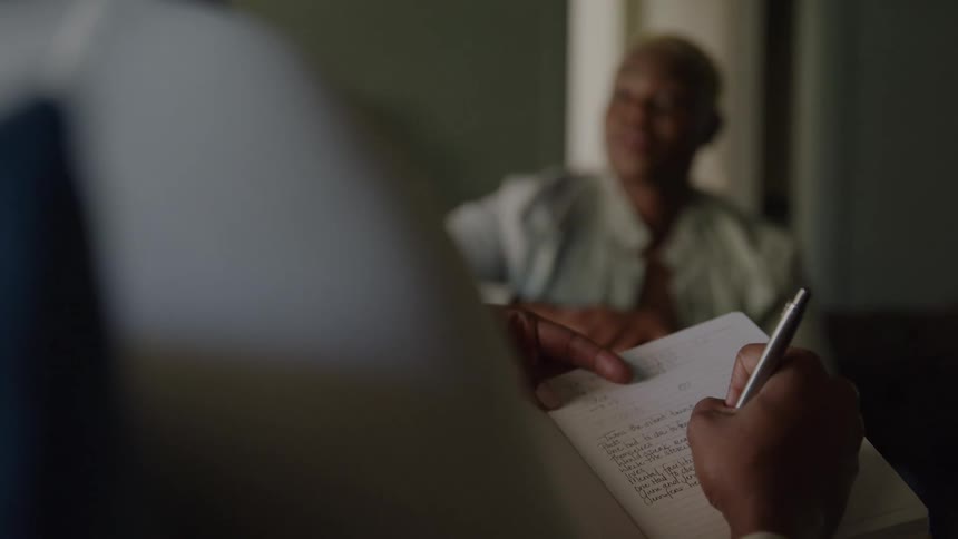 The video shows a closeup overtheshoulder view of someone writing notes in a notebook with a pen the writing is partially legiblein the background out of focus sits an older bald individual possibly a woman who appears to be talking or smilin