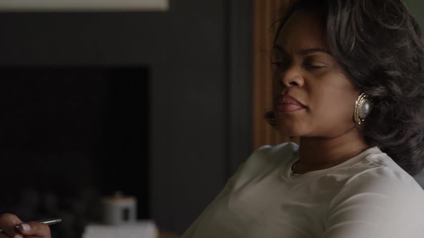 A closeup of an african american woman wearing a white top and a large decorative earring shows her looking intently to the side as she speaks with a serious or concerned expression she is holding a pen and looking down as she starts to make a note su