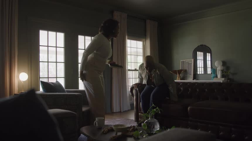 Two black adult females after walking and sitting on furniture are having a conversation in a dimly lit elegantly decorated living room with large windows