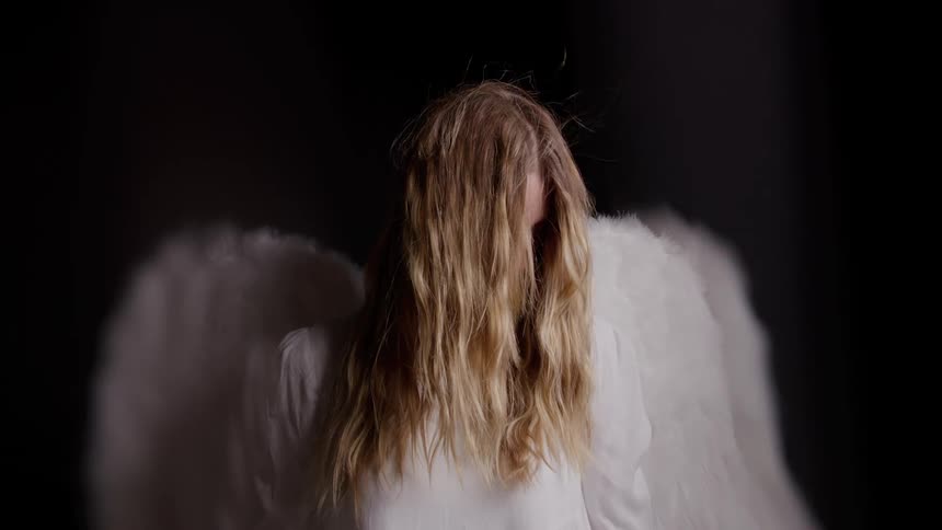 Woman with white decorative wings stands with head lowered wearing a lightcolored dress