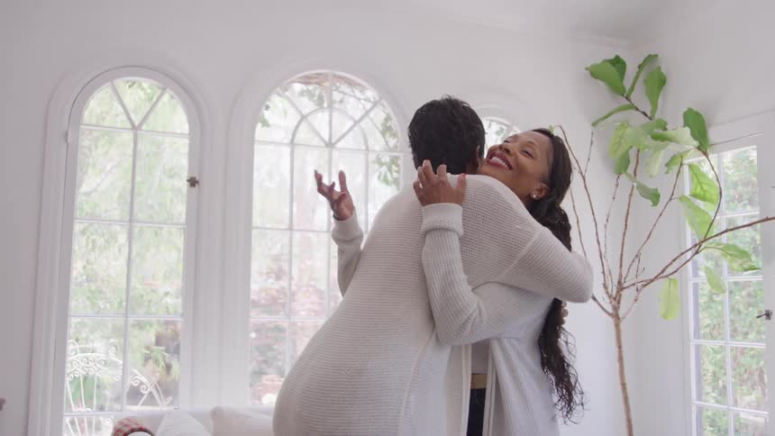 Two people hug in a bright room with tall windows in the background
