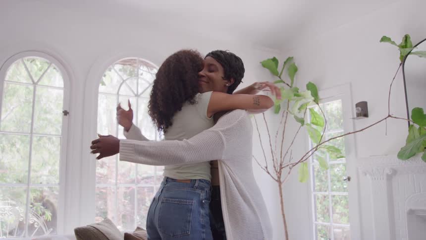 Two people hug in a bright room with large windows in the background