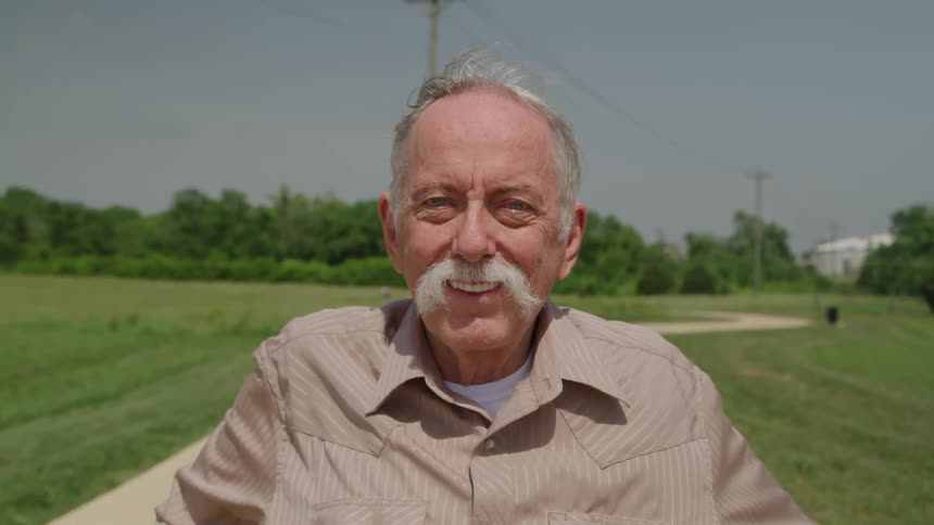 An older man with a grey mustache and hair smiles directly at the camera while wearing a tan collared shirt outdoors on a paved path in a grassy open park area on a clear day