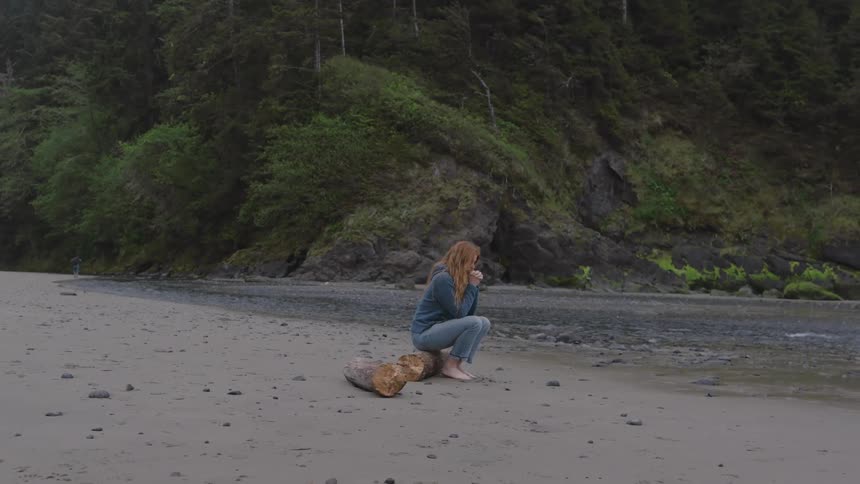 Woman sits on beach log gazing at the sea with hands under chin surrounded by nature